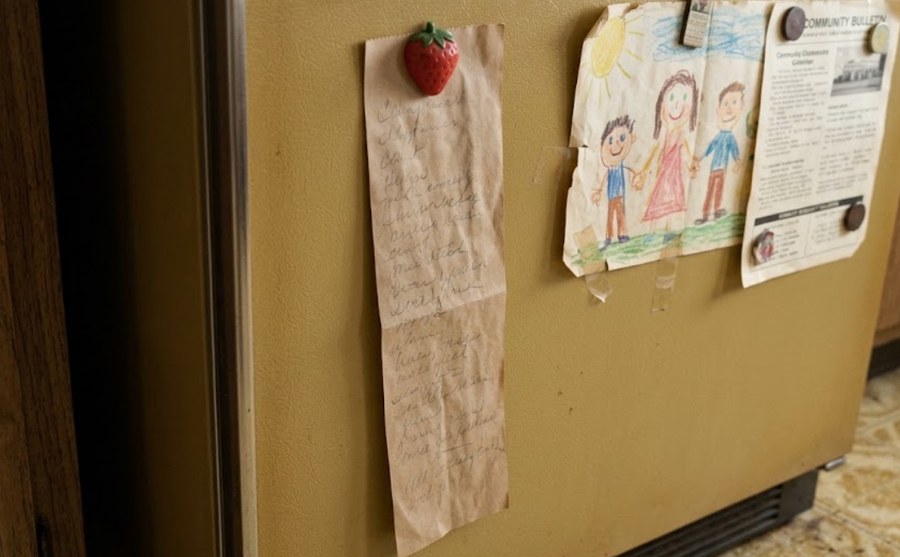 A paper bag grocery list taped to the refrigerator door with a strawberry magnet